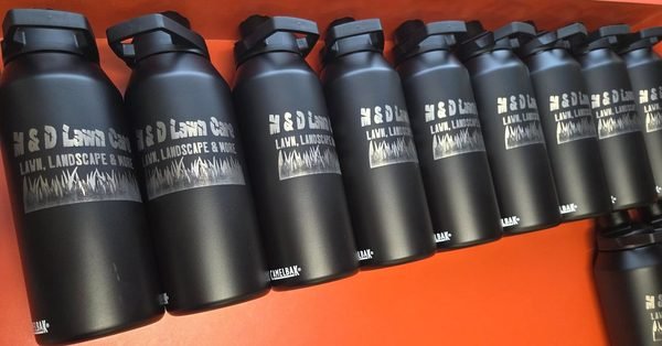 A row of black insulated bottles, each engraved with the H&amp;D Lawn Care, Landscape and More logo.