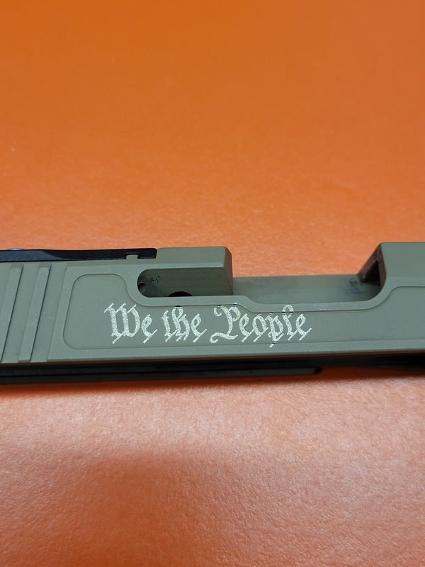 An olive drab pistol slide fiber-engraved in blackletter script with the words We the People.