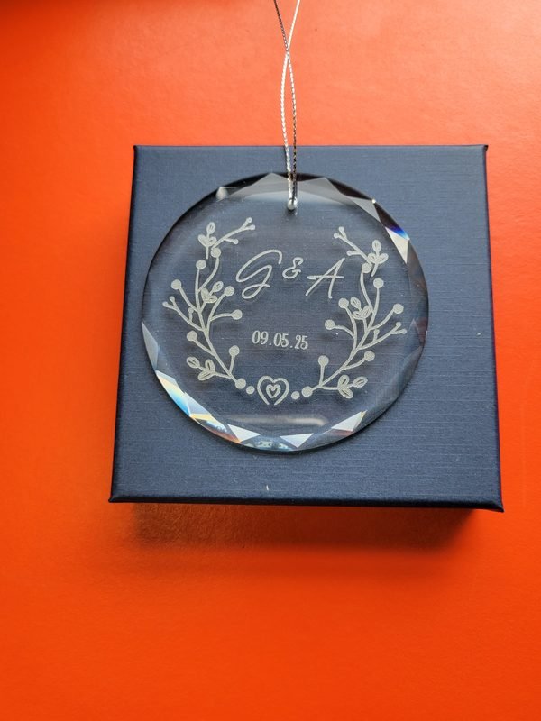 An engraved glass wedding ornament hanging on its ribbon above a navy presentation box.