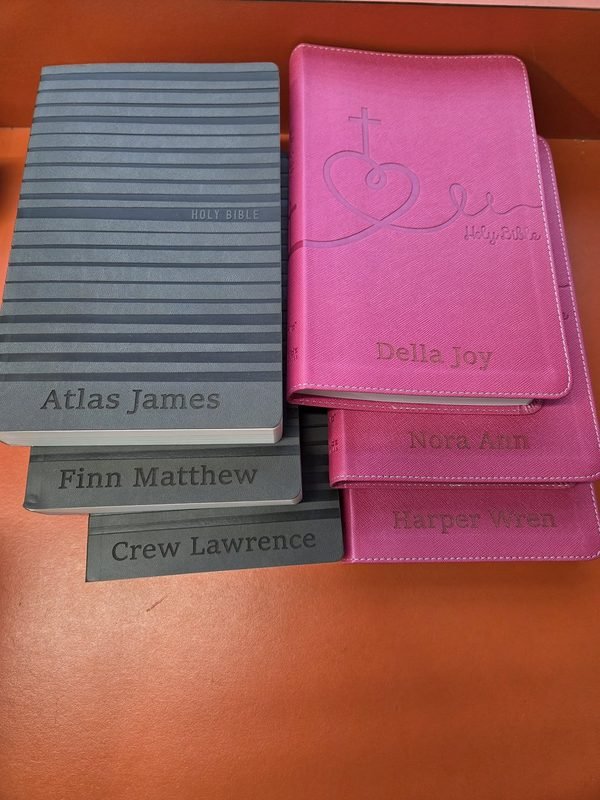 A stack of leather-bound Bibles in grey and pink, each engraved with a child's full name.