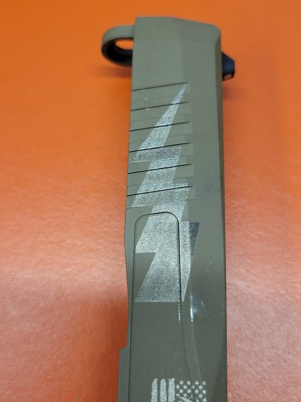 An olive drab pistol slide fiber-engraved with a lightning bolt and a United States flag.