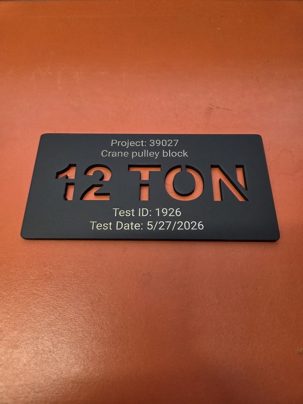 A black anodised tag with 12 TON cut clean through and Project 39027, Crane pulley block engraved beside it.