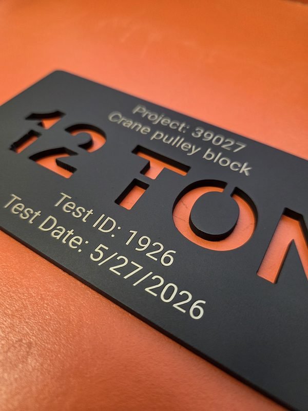 A close-up of a cut-through 12 TON load tag, the engraved test ID and date running along its edge.
