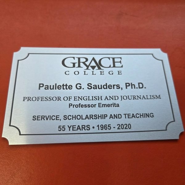 A brushed metal plaque with a fine engraved border, honouring a professor emerita's 55 years at Grace College.