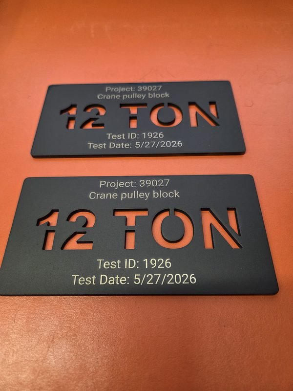 Two black anodised crane load tags with 12 TON cut through the plate and the test details engraved below.