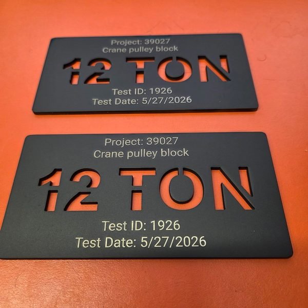 Two black crane load tags photographed from above, 12 TON cut through each plate.