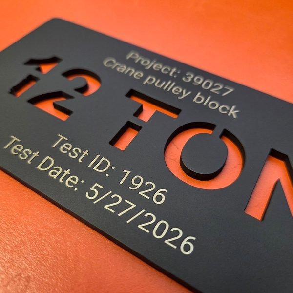 A sharply angled close-up of a black crane load tag, the cut lettering and engraved test date filling the frame.