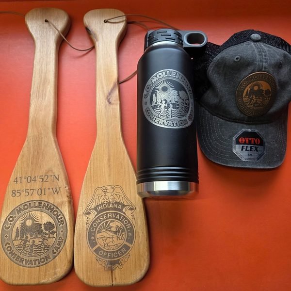Two engraved canoe paddles laid out with a matching camp bottle and leather-patch cap.