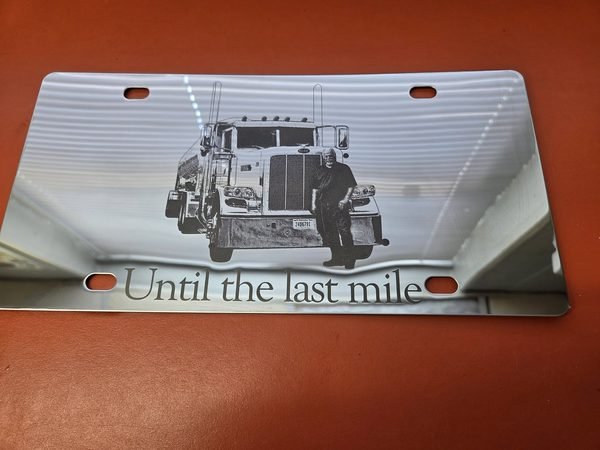 A brushed metal licence plate engraved with a driver leaning against his semi truck above the words Until the last mile.