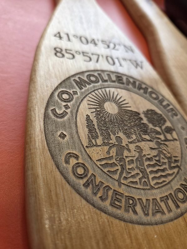 A close-up of a canoe paddle engraved with the C.O. Mollenhour Conservation Camp crest and the camp's coordinates.