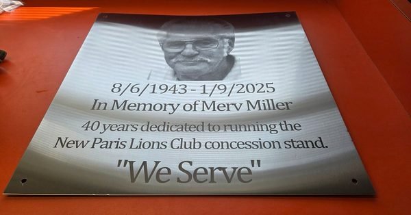A brushed metal memorial plaque engraved with a portrait of Merv Miller and forty years of service to the New Paris Lions Club.
