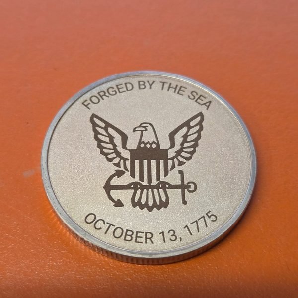 A challenge coin fiber-engraved with the Navy eagle and anchor above the words Forged By The Sea, October 13, 1775.