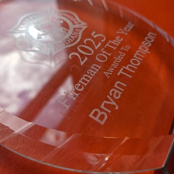 A close-up of a clear acrylic award, the Fireman Of The Year engraving frosted white against the light.