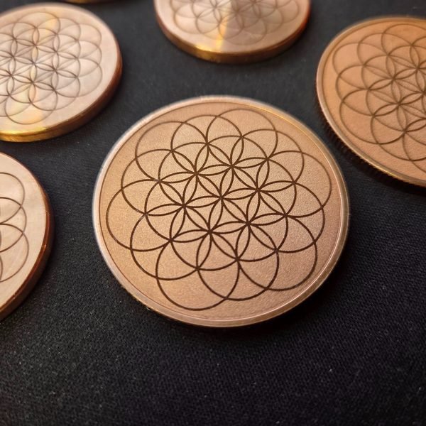 Copper discs on a dark cloth, each fiber-engraved with a fine Flower of Life pattern.