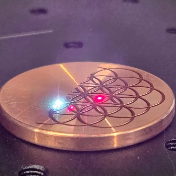 A copper disc on the laser bed mid-job, sparks flaring as the beam traces a geometric pattern.