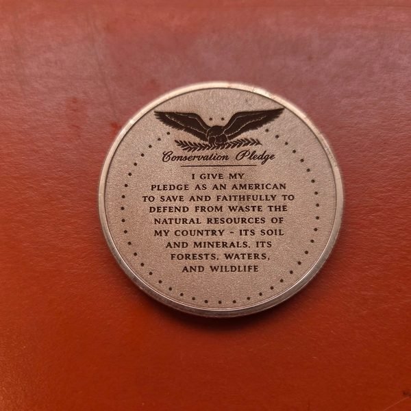 The reverse of a copper coin, fiber-engraved with the full text of the Conservation Pledge beneath an eagle.