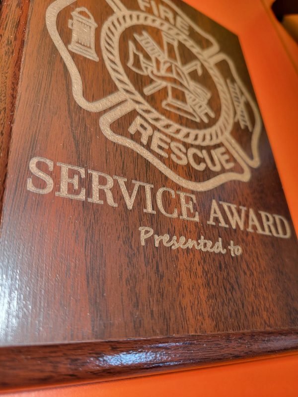A close-up of a wood-grain service award plaque, the fire rescue crest and lettering engraved deep into the surface.