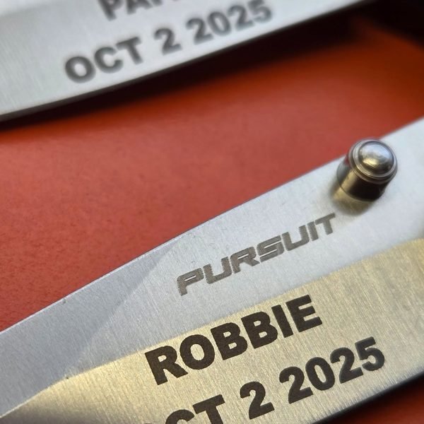 A macro view of a knife blade fiber-engraved ROBBIE, Oct 2 2025 beneath the Pursuit maker's mark.