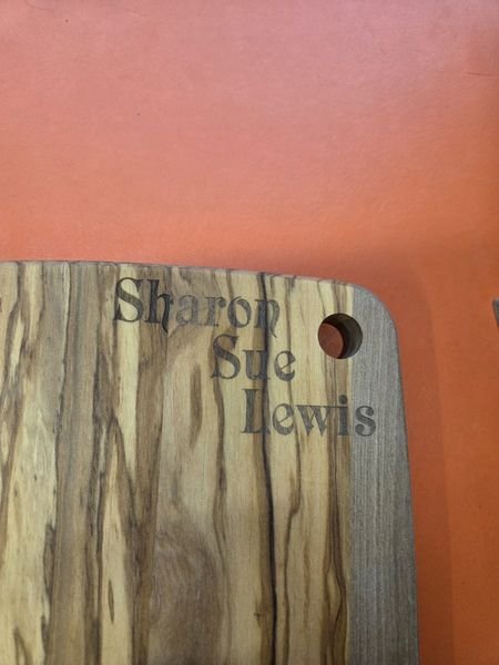 The corner of a marbled olive-wood cutting board engraved with the name Sharon Sue Lewis.