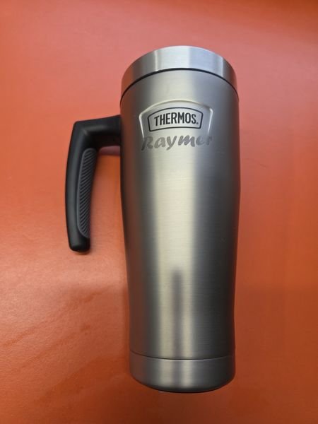 A stainless steel Thermos travel mug with a handle, engraved with the name Raymer beneath the maker's badge.