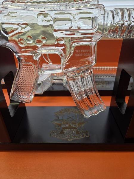 A rifle-shaped glass decanter etched 1LT Poppe, 2022-2026, on a black stand engraved with a unit crest.