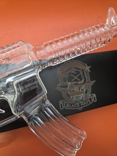 A rifle-shaped glass decanter etched SSG Guevara, 2022-2026, beside its stand engraved with the A Co 293rd Black Hats crest.