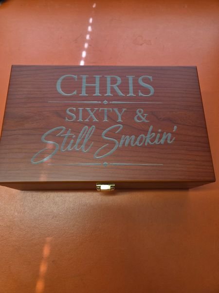 A wooden cigar box engraved Chris, Sixty & Still Smokin' in serif capitals and flowing script.