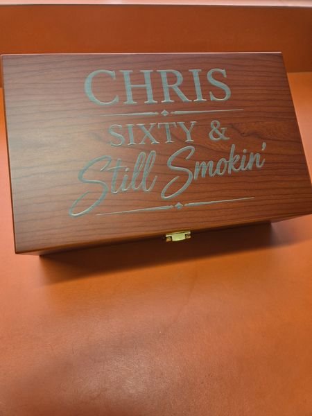 An angled view of the Chris, Sixty & Still Smokin' cigar box lid, the engraving pale against the dark wood grain.