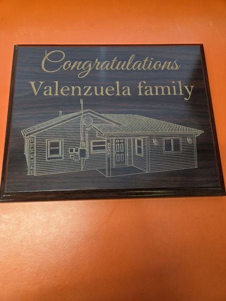 A dark woodgrain plaque engraved in gold with a congratulations message to a family above a line drawing of their house.