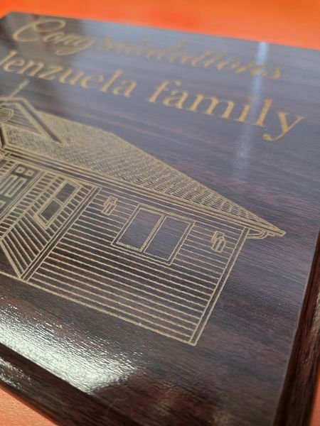 An angled close-up of the new-home plaque, the house drawing's siding, windows and shingles picked out in fine gold lines.