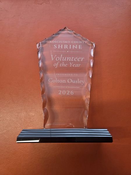 A clear crystal award with chiselled edges, on a black base, engraved Kosciusko County Shrine, Volunteer of the Year, 2026, with the recipient's name.