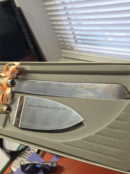 A wedding cake knife and server set in a grey presentation case, both blades engraved with the couple's names and wedding date in flowing script.