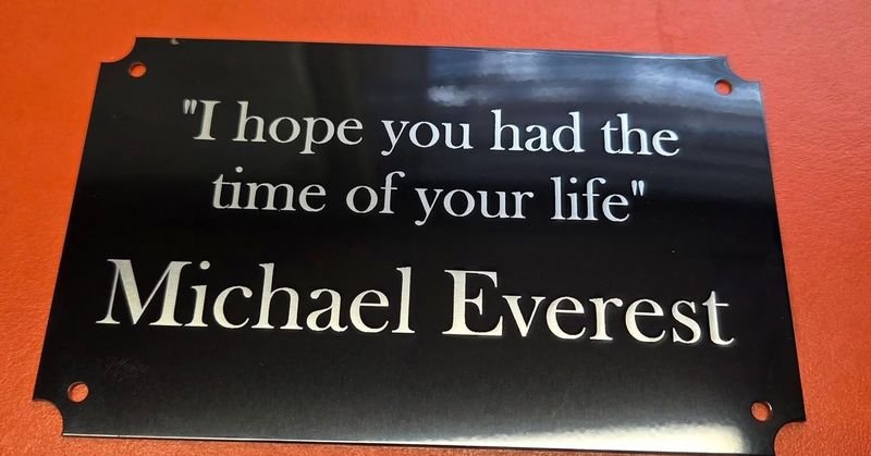 A black plaque engraved in bright serif lettering with a memorial line and a name.