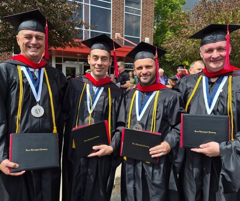 Four graduates holding diploma folders, each wearing an engraved medallion on a ribbon.