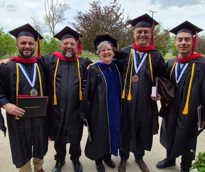 Five graduates in caps and gowns outdoors, wearing the medallions on ribbons.
