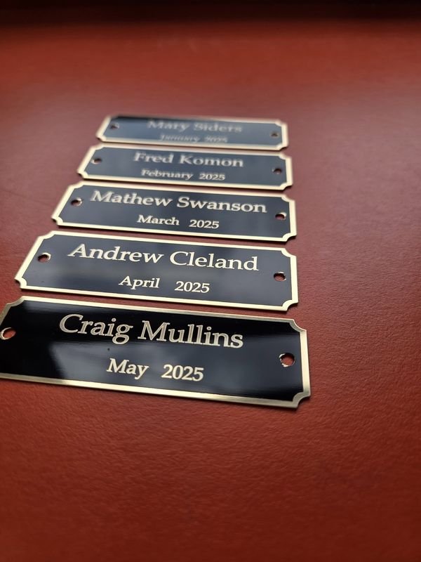 Five black-and-gold engraved name plates in a row, each with a name and a month of 2025.