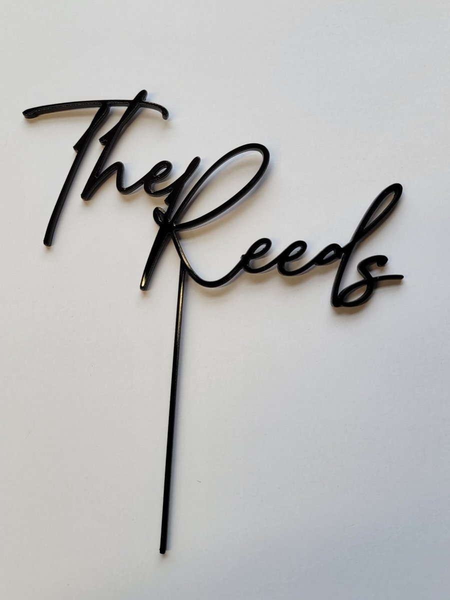 A black acrylic cake topper cut and engraved in flowing script reading The Reeds.